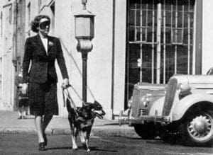 A black and white picture of a woman with a blind fold on being guided across a road by a German Shepard, she is wearing a blazer and knee length skirt and the fog has a short lead so is at her heal.