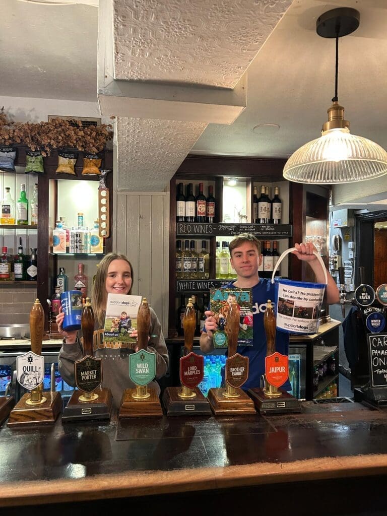 Paws for a Cause members Harriet and Ben behind the bar at The Stag's Head, holding donation buckets and leaflets for Support Dogs.