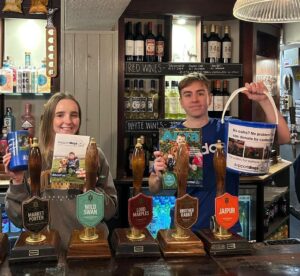 Paws for a Cause members Harriet and Ben behind the bar at The Stag's Head, holding donation buckets and leaflets for Support Dogs.