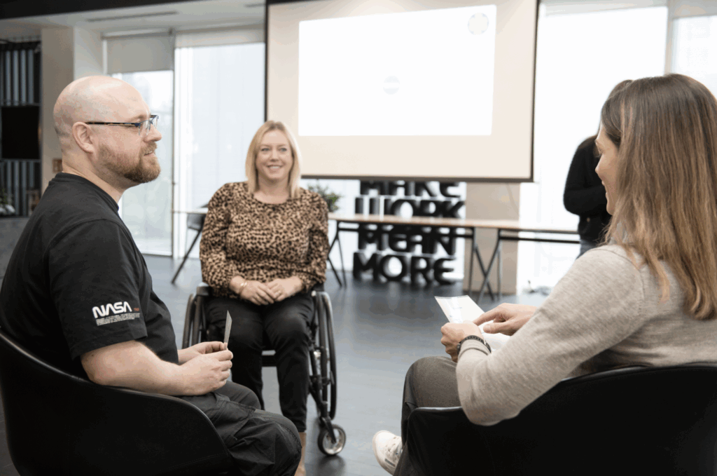 3 people sitting in a circle. They are all either seated or in a wheelchair. Woman at centre of image is wearing a patterned shirt.