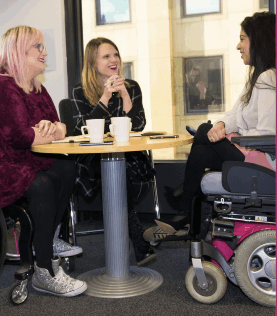 Three women, one in a wheelchair, speaking over coffee. They are all sat around a brown, round table.