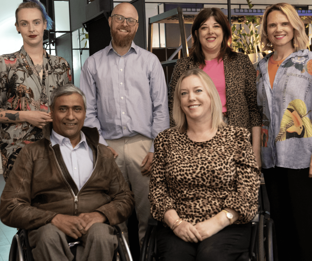 Group of men and women smiling for a picture. Four people in the back standing, two at the front in wheelchairs.